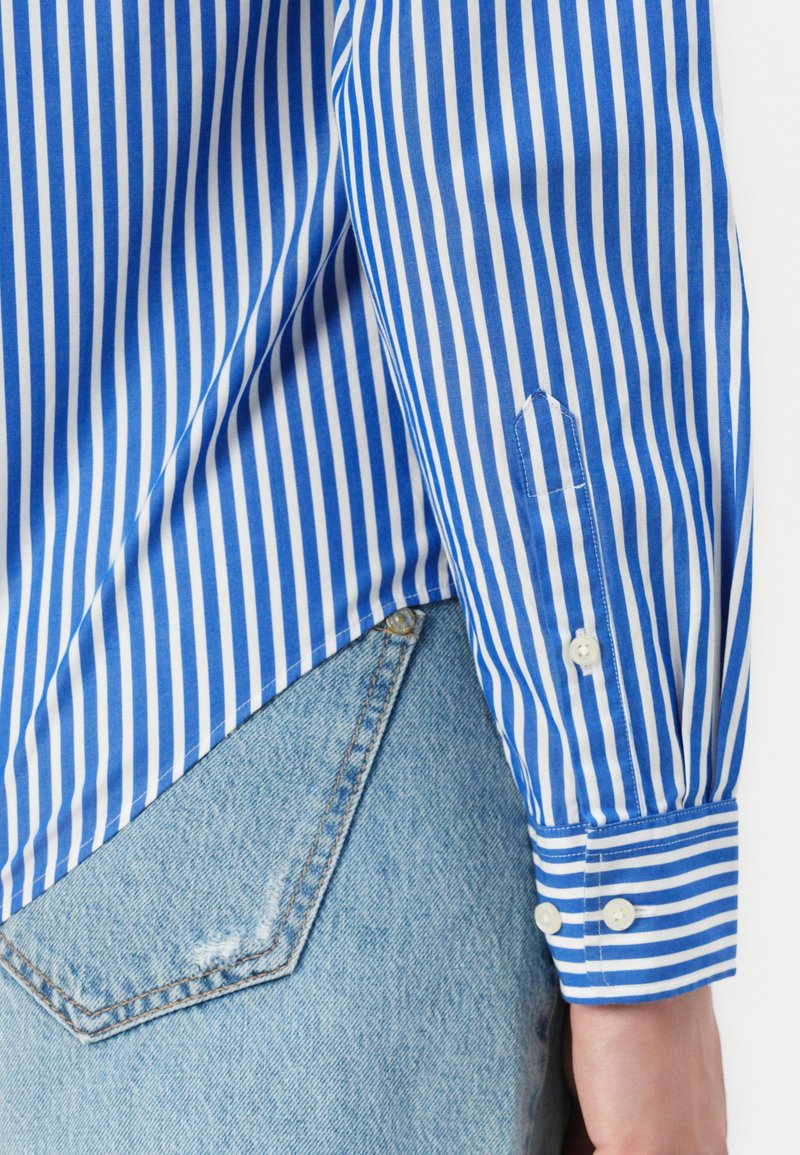 Blau-weiß gestreiftes Hemd mit Knopfmanschetten, mit spitzem Saum und leichtem Stoff, kombiniert mit Jeans.