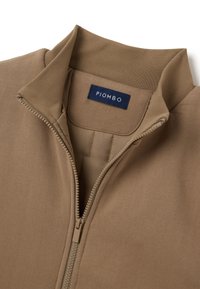 Beige Zip-Jacke mit hohem Kragen, die eine glatte Textur und leichten Stoff aufweist. "PIOMBO"-Etikett in Dunkelblau am inneren Kragen.
