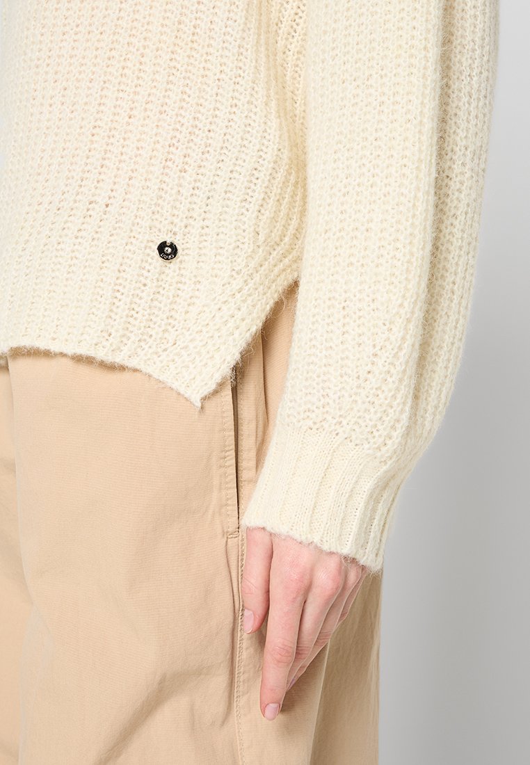 Crème-kleurige gebreide trui met een gestructureerd patroon, geribde manchetten en een kleine zwarte accentknop aan de zoom, vergezeld van beige broeken.