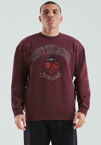 Mężczyzna z piegami ubrany w burgundowy sweter z napisem "RUTHLESS" i logo koszykówki, stojący na jednolitym jasnym tle.