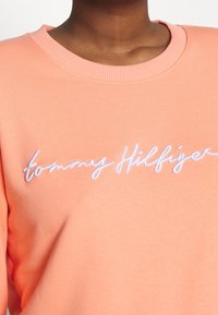 Roze sweater met geribde halslijn, voorzien van geborduurde witte "tommy Hilfiger" tekst op de borst. Zachte stoftextuur.