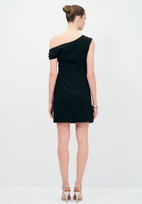 Schwarzes, tailliertes Kleid mit einseitigem Design, kurzer Länge und strukturiertem Stoff. Das Model trägt schlichte, metallische Accessoires und sandalenartige Absätze.