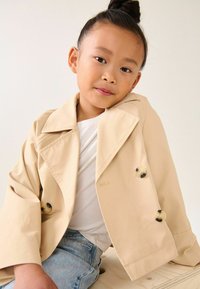 Beige trenchcoat met een brede kraag en twee grote, gemusterde knopen, gedragen over een witte blouse en lichtblauwe jeans. Glad materiaal.