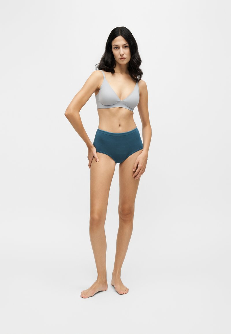 Femme debout pieds nus portant un soutien-gorge gris clair et un sous-vêtement taille haute bleu sarcelle foncé sur un fond clair uni.