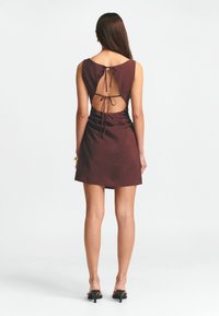 Mini abito di un intenso bordeaux con scollatura rotonda, schiena scoperta e dettaglio con fiocco. Tessuto liscio con una silhouette aderente e una leggera lucentezza.