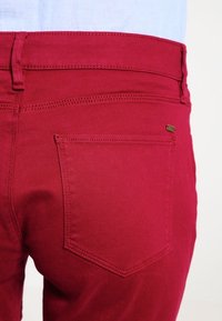 Vaqueros rojos de mezclilla con diseño clásico de cinco bolsillos, costuras visibles y acabado con herrajes metálicos. Tejido con textura y ligero brillo.