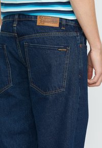 Jeans in denim blu scuro con taglio diritto, dotati di tasche posteriori, cuciture chiare e una patch in pelle con design impresso sulla cintura.