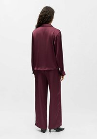Burgunderfarbene Seiden-Pyjama-Set mit einem locker sitzenden Hemd mit Kragen und langen Ärmeln, kombiniert mit weit geschnittenen Hosen und hohen Schuhen.