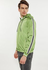 Giacca verde chiaro con zip e cappuccio, caratterizzata da accenti a righe bianche e nere lungo le maniche e una finitura lucida e strutturata.