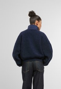 Mörkblå fleece-pullover med hög krage, med en texturerad yta och ribbade muddar. Matchas med mörka jeans.