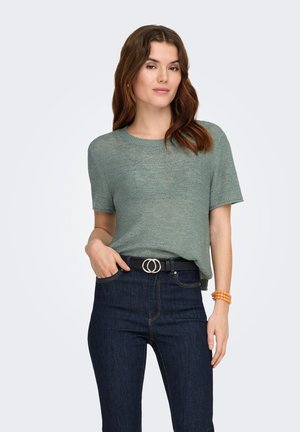 Kobieta o falowanych brązowych włosach, ubrana w zielony, krótki sweter z dzianiny, ciemnoniebieskie jeansy z czarnym paskiem z podwójną klamrą oraz pomarańczową bransoletkę.