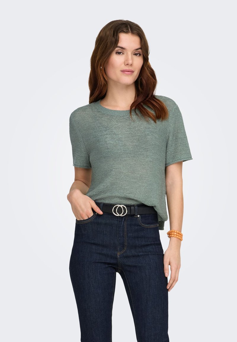 Frau mit welligem braunem Haar, die ein grünes, kurzärmeliges Strickoberteil, dunkelblaue Jeans mit einem schwarzen Gürtel mit Doppelring und ein orangenes Armband trägt.