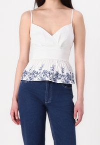 Haut péplum blanc avec broderie florale bleue à l'ourlet, doté de fines bretelles et d'un décolleté en V profond. Un jean en denim est porté en dessous.