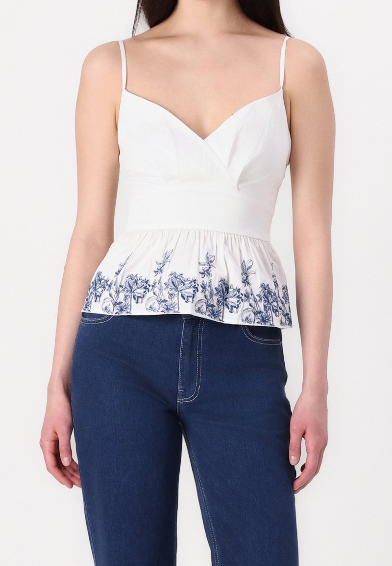 Haut péplum blanc avec broderie florale bleue à l'ourlet, doté de fines bretelles et d'un décolleté en V profond. Un jean en denim est porté en dessous.