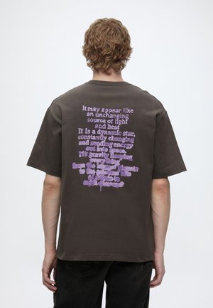 Person med krøllet hår set bagfra iført en mørkebrun T-shirt med lyse lilla tekst trykt på bagsiden.