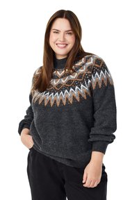 Žena se smiješi, nosi tamnosivi pulover s bijelim, narančastim i crnim geometrijskim uzorcima na prsima i rukavima, ruke u džepovima crnih hlača.