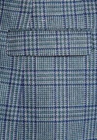 Tkanina tkana w zielono-niebieską kratkę, o fakturze, która wydaje się strukturalna i teksturowana. Zawiera kieszeń naszywaną.