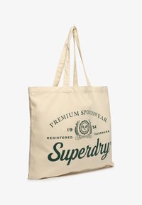 Beżowa torba na ramię wykonana z płótna, z zielonym tekstem zawierającym logo "Superdry" oraz dekoracyjnymi elementami. Dwie mocne rączki na ramię.