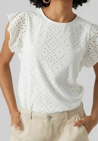 Donna con una blusa bianca in pizzo a occhiello con maniche a volantino e pantaloni beige a vita alta, mani in tasca.