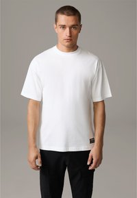 Weißes Baumwoll-T-Shirt mit Rundhalsausschnitt und kurzen Ärmeln. Verfügt über ein Etikett an der unteren Seite, kombiniert mit schwarzen Cargo-Hosen.