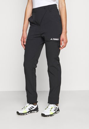 Osoba ubrana w czarne outdoorowe spodnie Adidas Terrex i białe buty do biegania trailowego Adidas na jednolitym tle.