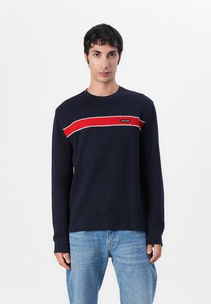 Marineblaues Langarm-T-Shirt mit geripptem Muster, das einen horizontalen roten Streifen und weiße Akzente aufweist, Logo-Patch auf der Brust. Kombiniert mit blauen Jeans.