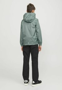 Giacca verde leggera con cappuccio, polsini ed orlo elasticizzati; abbinata a pantaloni neri e scarpe da ginnastica nere con dettagli bianchi.