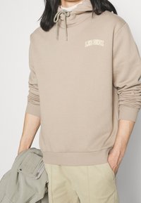Felpa beige con collo alto, chiusura con coulisse, polsini e orlo a costine, logo ricamato "LES DEUX" in giallo chiaro sul petto.