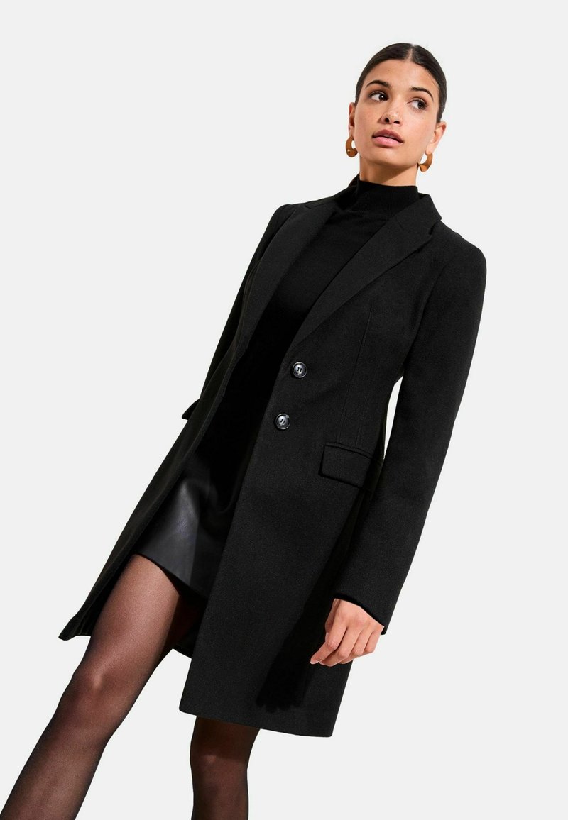 Fekete gyapjúkabát szabott fazonban, kiképzett nyakkal, két gombbal és oldalzsebekkel. Fekete garbó és szoknya fölött viselve.