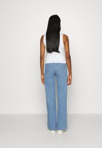 Jeans de denim azul claro, com corte flare e cintura alta. O tecido tem uma textura lisa. Apresenta dois bolsos traseiros e detalhes de costura sutis.