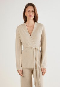 Suéter envolvente beige en material de punto suave, con mangas largas, un cinturón atado a la cintura y un escote en V profundo, con bordes acanalados.