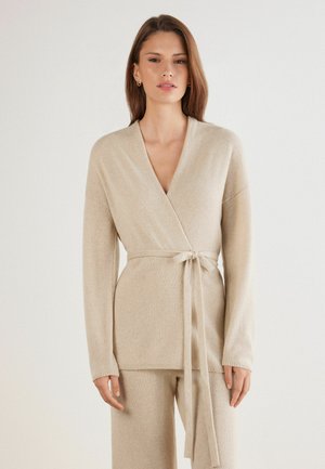 Pull wrap beige en maille douce, présentant des manches longues, une ceinture à nouer à la taille et un décolleté en V profond, avec des bords côtelés.