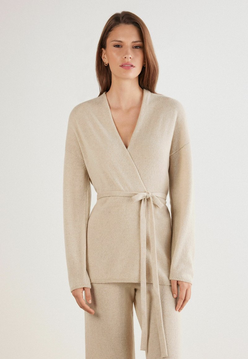 Suéter envolvente beige en material de punto suave, con mangas largas, un cinturón atado a la cintura y un escote en V profundo, con bordes acanalados.