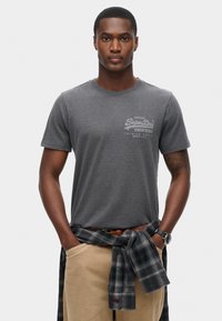 Grå bomulds T-shirt med rund hals, der har et diskret logo print på brystet. Sættes sammen med khaki bukser og en sort ternet skjorte bundet om livet.