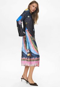 Largo vestido negro con motivos coloridos inspirados en el circo, con mangas acampanadas y una silueta fluida. Los zapatos son marrones con punta afilada.