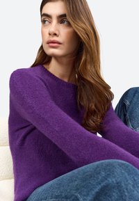 Purple knit sweater with a round neckline, soft texture, and long sleeves, paired with dark blue denim jeans. Model seated, neutral background.