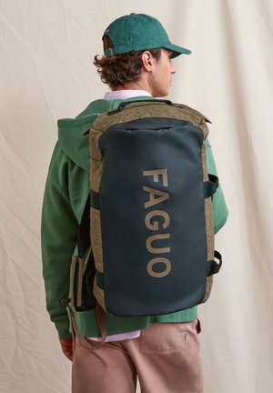 Homme portant un sweat à capuche vert et une casquette, portant un grand sac de sport kaki et noir avec le texte "FAGUO" au dos.