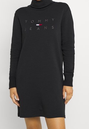Czarna sukienka z długim rękawem i golfem z logo Tommy Jeans oraz małym czerwono-biało-niebieskim emblematem flagi na piersi.