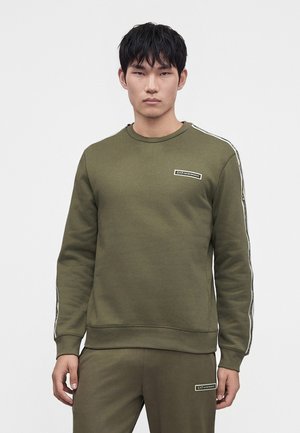 Jeune homme portant un sweat-shirt vert olive et un pantalon assorti avec des écussons du logo EA7 Emporio Armani sur la poitrine et la cuisse.