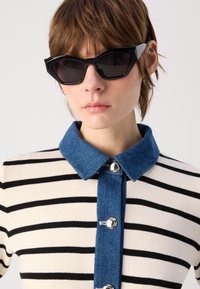 Occhiali da sole neri a forma di occhio di gatto con lenti scure. Il modello indossa una camicia a righe beige e nera con colletto in denim e bottoni argentati.