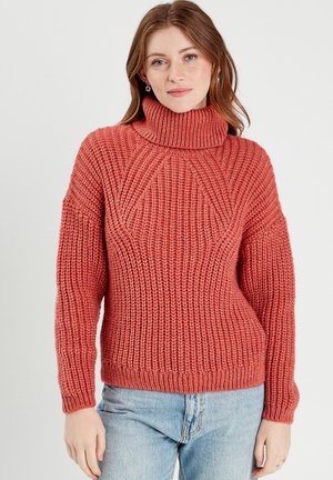 Rust-colored turtleneck sweater with a chunky, knitted texture, featuring a ribbed hem and drop shoulder design. Worn with light blue jeans.
