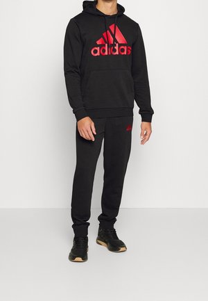 Homme portant un sweat à capuche noir Adidas avec un grand logo rouge, un pantalon de survêtement noir assorti Adidas et des baskets noires avec des semelles couleur sable.