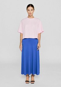 Helles rosa, kurzärmeliges Crop-Top, kombiniert mit einem langen blauen Rock. Das Outfit zeichnet sich durch eine glatte Textur und ein schlichtes Design aus. Schwarze Riemchensandalen.