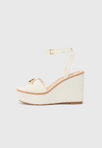 Sandales compensées en crème avec une semelle extérieure tissée texturée blanche, une bride de cheville réglable et un accent en métal doré sur les orteils.