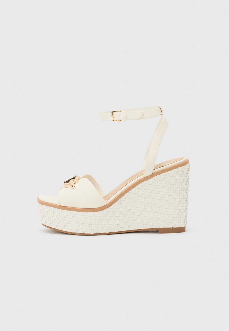Sandales compensées en crème avec une semelle extérieure tissée texturée blanche, une bride de cheville réglable et un accent en métal doré sur les orteils.