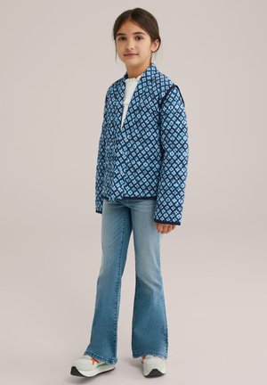 Vadderad blå jacka med geometriska mönster, i kombination med ljusblå utsvängda jeans och flerfärgade sneakers, vilket ger en avslappnad, lager-på-lager-look.