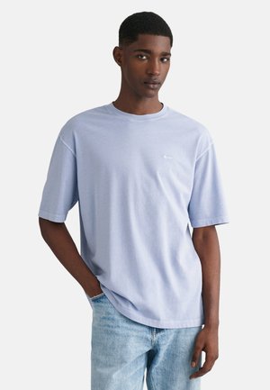 Camiseta de manga corta azul claro, hecha de algodón, con cuello redondo y un pequeño logo en el lado izquierdo del pecho. Combinada con jeans de mezclilla claros.