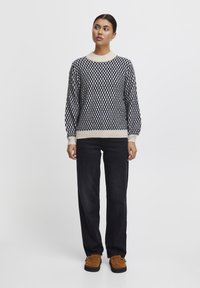 Maglione lavorato a maglia in blu navy e crema con motivo a diamante, collo rotondo, polsini e bordo a costine. Abbinato a pantaloni neri e mocassini marroni.