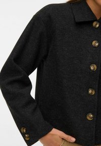 Veste en laine gris foncé avec de gros boutons en écaille de tortue sur le devant et deux boutons sur la manche, portée avec la main dans la poche.