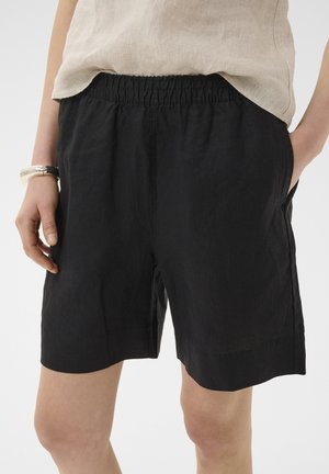 Nő fekete, rugalmas derekú, zsebes rövidnadrágot és bézs színű, lenvászon felsőt visel, kezei mellett áll, egyszínű háttér előtt.